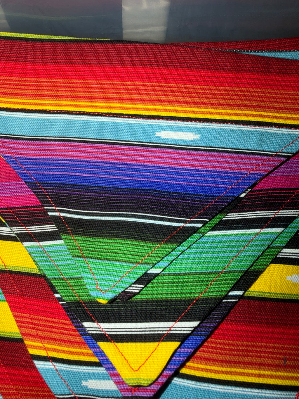Close-up of a colorful woven textile with horizontal and diagonal striped bands in red, yellow, orange, blue, green, black, white, and pink, featuring visible red stitching.