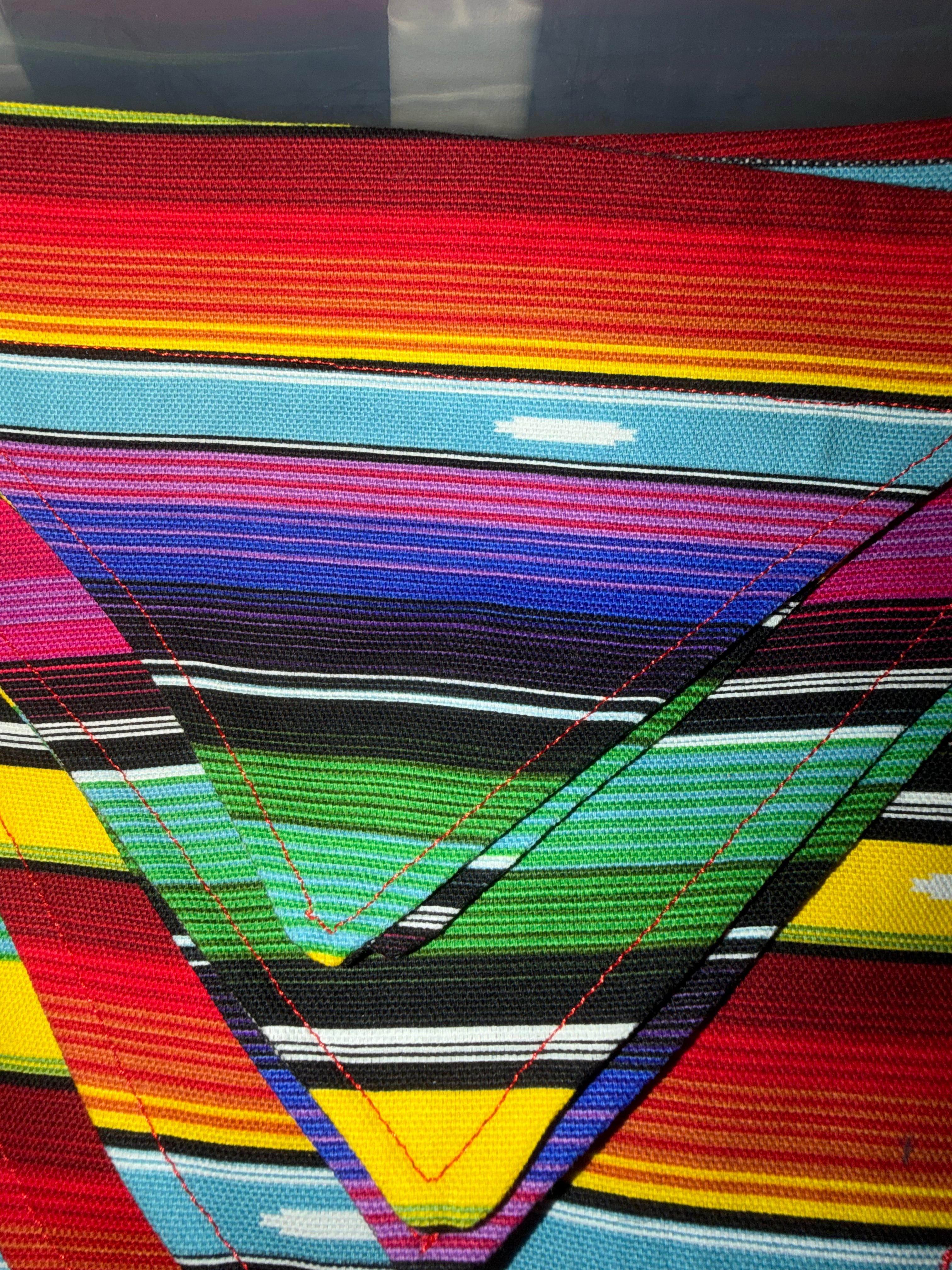 Close-up of a colorful woven textile with horizontal and diagonal striped bands in red, yellow, orange, blue, green, black, white, and pink, featuring visible red stitching.